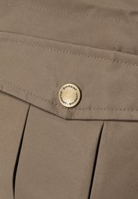 Beige auduma bikses ar priekšējiem krokām un metalīgu pogu ar iegravētu zīmolu. Tekstūra ir gluda, ar redzamām šuvju detaļām.