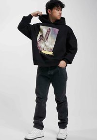 Svart huvtröja med grafiskt tryck av en rymdraketuppskjutning; har en känguruficka och ribbade ärmslut, i kombination med mörka jeans och vita sneakers.