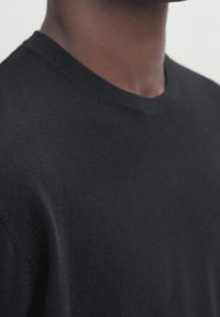 Gros plan sur une personne portant un pull noir uni à col rond, montrant une partie du cou et de l'épaule sur un fond clair.