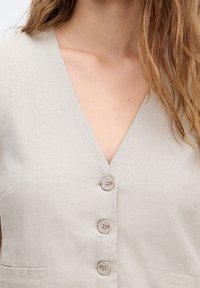 Jasno beżowa lniana kamizelka z dekoltem w serek i czterema dekoracyjnymi guzikami z przodu. Posiada usztywnioną strukturę oraz boczne kieszenie.