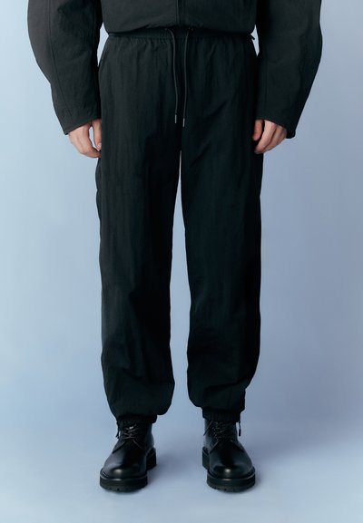 Black, lightweight trousers with a relaxed fit, elasticized ankles, and adjustable waistband. Worn with black lace-up boots.