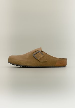 Sabot en daim marron à enfiler avec une bride à boucle réglable et une semelle noire rainurée, vue en angle sur fond neutre.