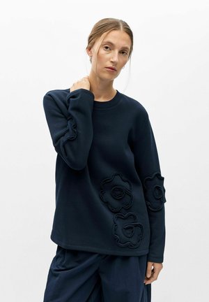 Femme aux cheveux châtain clair porte un sweat-shirt bleu marine avec des motifs floraux en relief sur les manches et le devant, associé à un pantalon bleu marine ample.