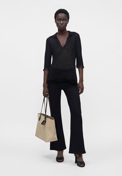 Femme vêtue de noir avec des manches trois-quarts et un pantalon évasé tenant un sac cabas beige avec des poignées foncées sur un fond uni.