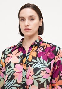 Chemise à boutons avec un motif floral, présentant des fleurs vibrantes roses, oranges et violettes sur fond noir. Fabriquée en tissu texturé léger.