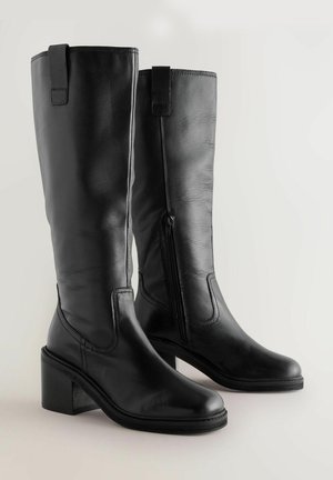 Botas de cuero negro hasta la rodilla con tacón cuadrado, con una cremallera lateral y una lengüeta para facilitar el uso. Textura suave y costura definida.