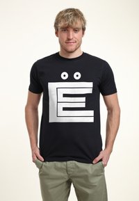 Svart t-shirt med kort ärm som har ett stort vitt blockbokstav "E"-motiv med tecknade ögon ovanför. Tillverkad av bomullstyg.
