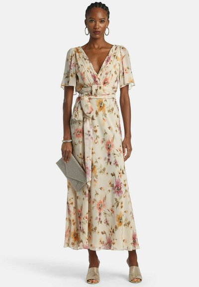 Nő krémszínű, virágmintás átlapolt ruhában, fodros ujjakkal, öves derékkal, texturált borítéktáskával a kezében, nyitott orrú cipőben és karikafülbevalóban.