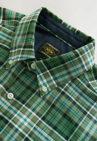 Grøn- og blåternet skjorte med button-down krave, der har et mærke med teksten "NEXT" og to hvide knapper foran.