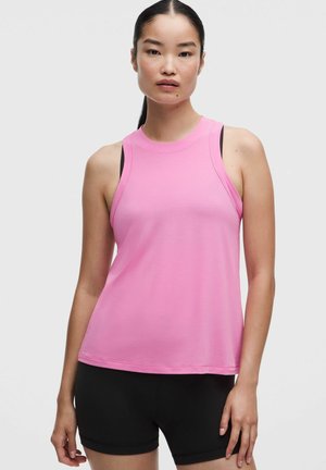 Giovane donna con lunghi capelli scuri indossa un top rosa senza maniche sopra una canotta nera e pantaloncini neri su uno sfondo chiaro uniforme.