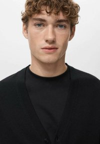 Czarny kardigan z dekoltem w serek, o gładkiej fakturze i zapinany na guziki. Noszony na czarnej koszulce, pokazany na jednolitym tle.