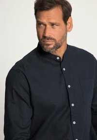 Námořnická modrá košile s dlouhým rukávem z texturované látky, s krkem na pevno, přední kapsou a světlými knoflíky na jednobarevném pozadí.