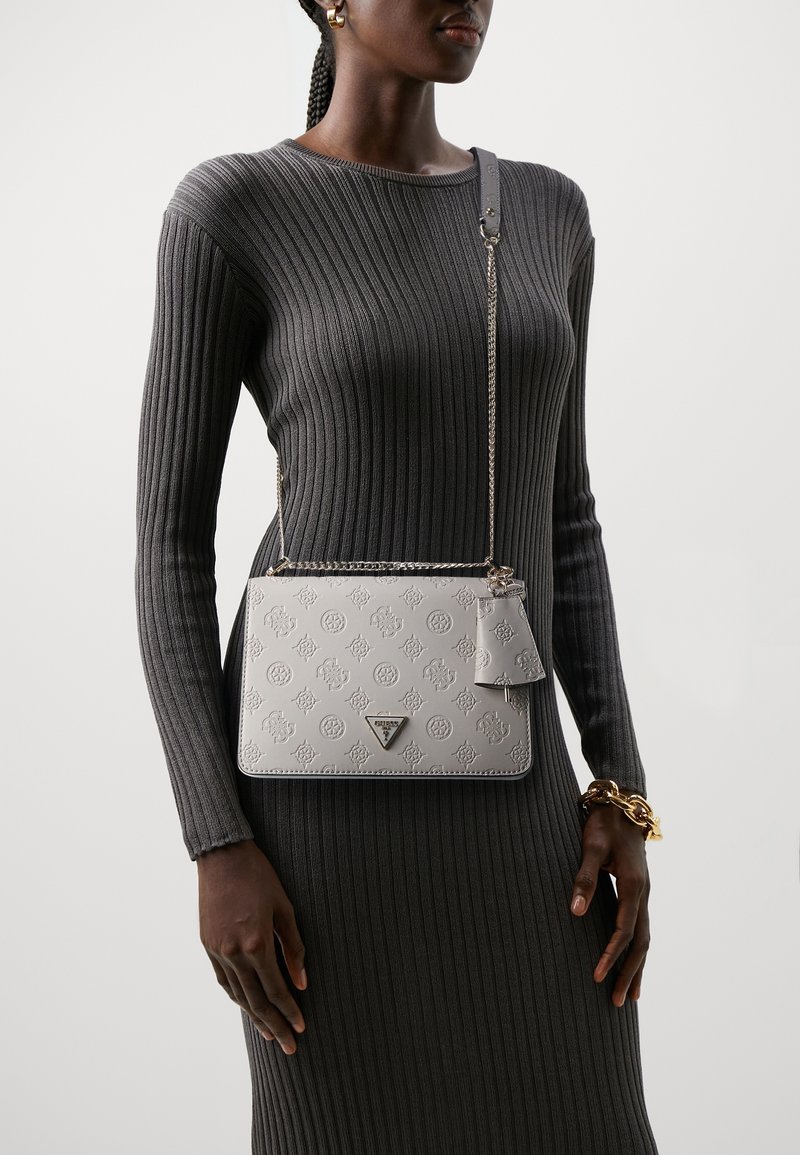 Femme portant une robe côtelée grise avec un sac bandoulière blanc embossé Guess et des bijoux dorés sur un fond uni.