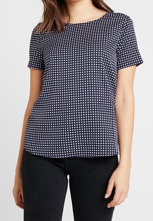 Woman wearing a navy blue short-sleeve top with white square patterns paired with black pants, standing against a plain background.