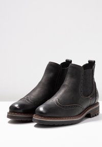 Černé kožené kotníkové boty s elastickými bočními panely, dekorativní perforací brogue, zaoblenou špičkou a texturovanou podrážkou. Kontrastní prošití.