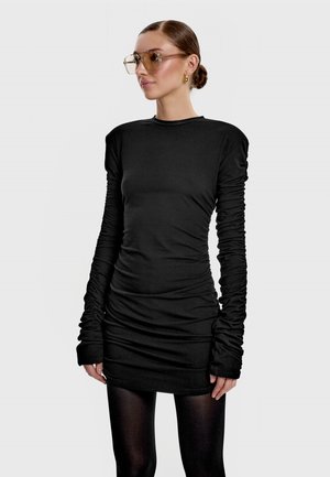 Kobieta z włosami związanymi w kok, nosząca złote kolczyki koła, oversize'owe przyciemniane okulary oraz czarną mini sukienkę z marszczeniami i długimi rękawami oraz czarne rajstopy.