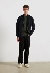 Chaqueta bomber de lana azul con cremallera sobre una camisa a rayas verdes, combinada con pantalones negros y zapatillas beige. La chaqueta presenta un logo discreto.