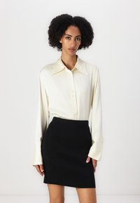 Cămașă din satin crem, cu mâneci lungi și guler ascuțit, asociată cu o fustă neagră cu textură ribbed. Cămașa are detalii subtile cu nasturi.