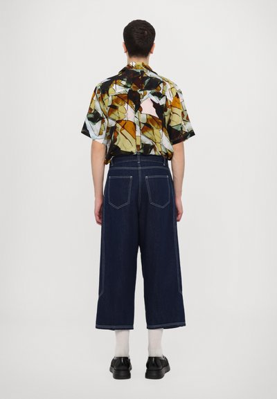 Kortærmet skjorte med et farverigt abstrakt mønster; mørk denim bukser med vidde og kontraststing; sorte sko. Hvid baggrund.