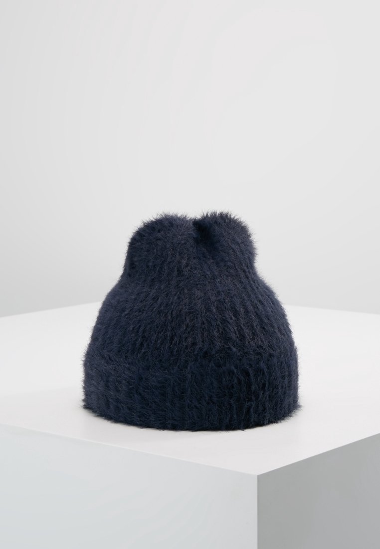 Bonnet tricoté bleu marine foncé en maille duveteuse présenté sur un piédestal blanc contre un fond gris clair uni.
