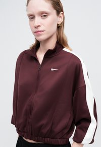 Burgundská zipová bunda s bielymi bočnými pruhmi, elastickým pásom a logom Nike. Vyrobená z hladkej tkaniny, s vysokým golierom a krátkymi rukávmi.