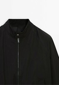 Veste légère noire avec fermeture éclair frontale, col à boutons-pression et détails de couture diagonale sur la zone des épaules.