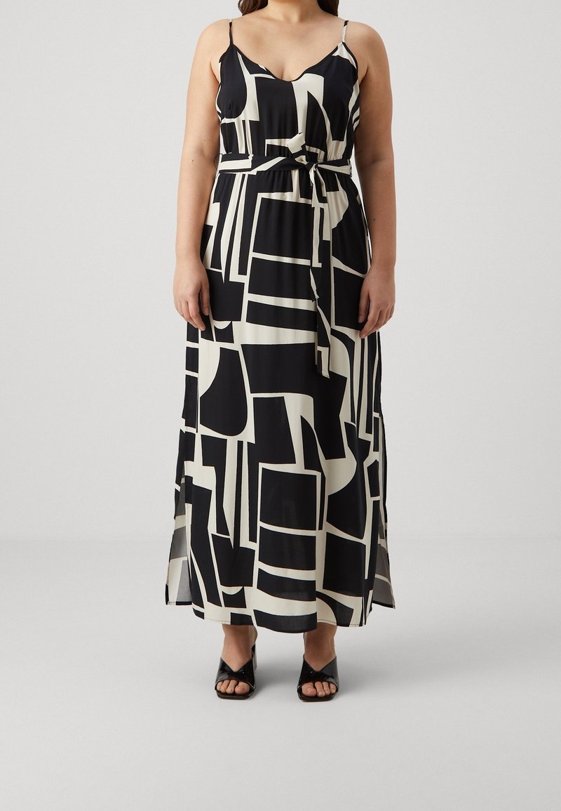 Femme portant une robe maxi sans manches à motif géométrique noir et blanc, taille nouée et talons ouverts noirs.
