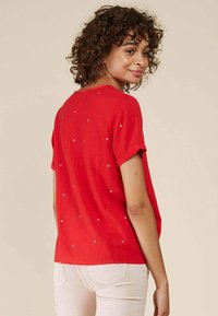 Chemise rouge à manches courtes avec une coupe décontractée, présentant un motif texturé de petits triangles métalliques dispersés au dos.