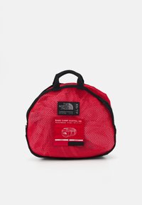 Red mesh duffel bag with black accents. Features a rectangular shape, dual handles, and a label with material and dimensions.