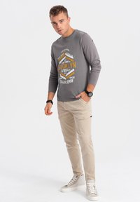 Grau langärmliges T-Shirt mit grafischem Druck, kombiniert mit hellbeigen Cargo-Hosen. Das Outfit wird mit weißen Sneakers und einer schwarzen Armbanduhr abgerundet.