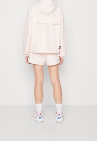 Jasnoróżowa wiatrówka z kapturem, elastycznymi mankietami i tylną kieszenią. W komplecie z pasującymi szortami. Białe i czerwone buty sportowe z niebieskimi akcentami.