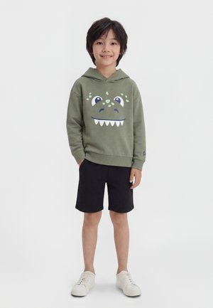 Băiat tânăr în picioare, purtând un hanorac verde cu fața unui dinozaur desen animat, pantaloni scurți negri și adidași albi, zâmbind către cameră.