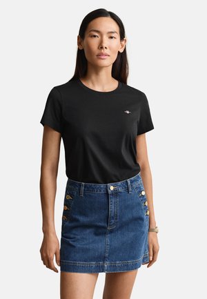 Woman wearing a black t-shirt with a small logo and a blue denim skirt featuring gold buttons on the front sides.