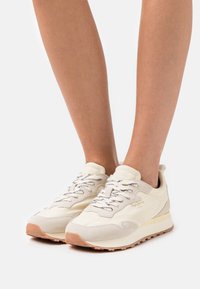 Nohy obuté v krémových a béžových teniskách Pepe Jeans s bílými a černými tkaničkami, stojící na bílém pozadí.