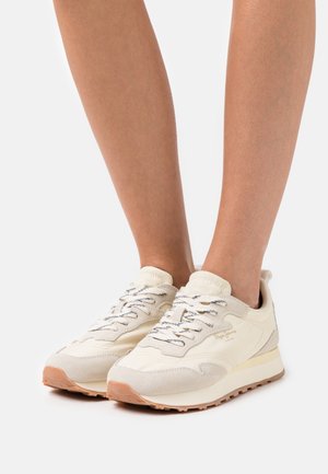 Nohy obuté v krémových a béžových teniskách Pepe Jeans s bílými a černými tkaničkami, stojící na bílém pozadí.