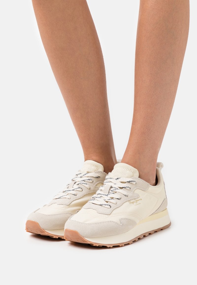 Nohy obuté v krémových a béžových teniskách Pepe Jeans s bílými a černými tkaničkami, stojící na bílém pozadí.