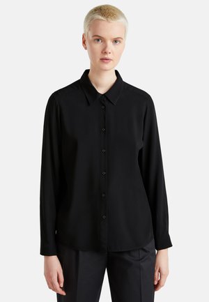Personne aux cheveux blonds courts portant une chemise noire à manches longues boutonnée et un pantalon noir sur un fond clair uni.