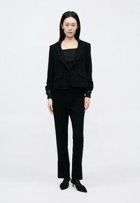 Sieviete stāv, valkājot melnu žaketi ar sporta kapuces auklām, atbilstoša melna krāsā bikses un apavus ar smailu purngalu, pret vienkāršu balto fonu.