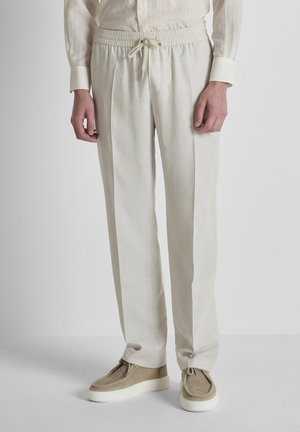 Hombre con pantalones beige con cordón, zapatos casuales beige y una camisa cremosa texturizada, de pie contra un fondo claro y liso.