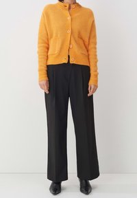 Stickad orange kofta med rund hals, fem knappar och två framfickor. Kombinerad med hög midja svarta byxor och svarta skor.