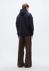 Φούτερ με κουκούλα σε ναυτικό μπλε, με τσέπη τύπου καγκουρό και ρυθμιζόμενο κορδόνι στην κουκούλα, συνδυασμένο με καφέ παντελόνια φαρδιάς γραμμής με τσέπες και υφή.