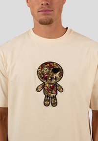 Beige Baumwoll-T-Shirt mit einem detaillierten gestickten Grafikdesign einer Puppe, verziert mit Mustern in Gold, Rot und Schwarz, einschließlich Herz- und Knopfakzenten.