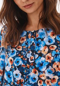 Blusa floreale con stampe blu e arancioni su sfondo nero, dotata di scollo rotondo e dettagli con bottoni. Tessuto morbido e leggero.