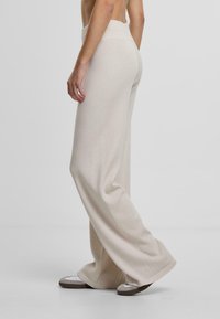 Pantalons larges beige en tissu tricoté texturé, avec une taille haute et une silhouette allongée, assortis à des chaussures grises à enfiler.