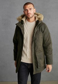 Olivově zelená parka s kapucí lemovanou umělou kožešinou, se zapínáním na knoflíky a zip, a několika kapsami, nošena přes krémový svetr s pleteným vzorem.