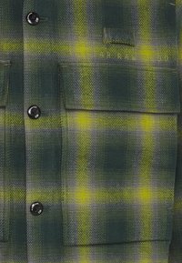 Grön och gul rutiga flanellskjorta med svarta knappar och två bröstfickor, med en texturerad tyg och synlig varumärkeslogotyp.