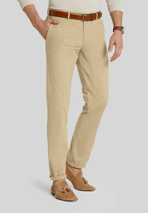 Man wearing beige trousers, brown leather belt, watch, and tan suede loafers with tassels, standing with one hand in pocket.