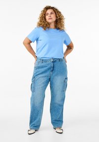 T-shirt in cotone azzurro chiaro abbinata a jeans cargo a lavaggio chiaro con vestibilità morbida, dotati di tasche laterali e forma rilassata. Scarpe a motivo bianco e nero.