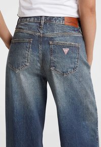 Spijkerbroek met een vervaagde blauwe was, klassiek vijf-pocket ontwerp en een driehoekige rode logopatch op de achterzak.