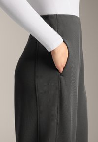 Nohavice z rebrovanej tmavosivej tkaniny majú vysoký pás a bočné vrecká. Dlhý rukáv bieleho topu kontrastuje s nohavicami.